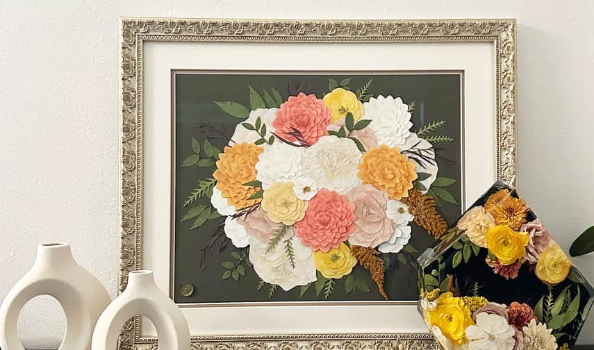 Pressed wedding bouquet in an ornate gold frame, featuring coral, blush, white, and yellow flowers on a deep green background.